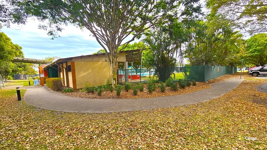 A concrete pathway with a rough surface connects the car park to a low-set cream brick building with light brown brick foundation edges. Black safety bollards can be seen on the side of the concrete pathway. Covering the landscape is dried leaf litter. Small green plants with parallel leaf venation can be seen on the right, just before the building.