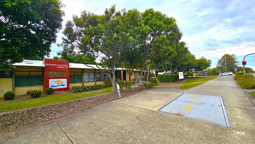 Closer look leading to another entry point closer to the main highway. Image shows several low-set small cream brick buildings with glass windows that have green outline frames and light brown brick foundation edges. Seen from this position is a large red wall cover detailing the Murrumba Community Swimming Pool on top and the logo on a white background below. Shrubs and bushes can be seen against the walls of the buildings.