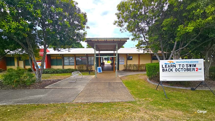 Entry to Murrumba Downs Swimming Pool. On the right side is a white signboard with the name of the facility and the light brown and blue logo. A ‘Learn to Swim Back October 2nd’ can be read on the white sign board. At the left side of the entrance is a stainless steel bike rack. There is a tall entry porch with lime green metal frames and corrugated roofing that connects to the corridor inside the entire facility.