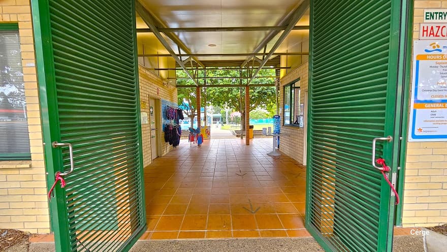 Underneath the tall entry porch with lime green metal beams and corrugated roofing of Murrumba Downs Swimming Pool. The other end of the entry porch is supported by a light brown brick foundation. Place against the left light brown brick foundation of the porch is a light blue signboard. Entrance is through green steel paneled Louvre doors right after the porch. Glass windows are noticeable on the left side.