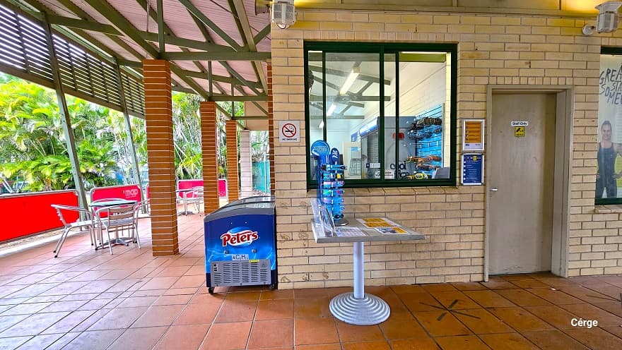 Reception and retail at Murrumba Downs Swimming. On the right side of the entrance is the ‘Staff Only’ brown door with a yellow CCTV sign. Next to the door is a glass window with green metal frames. In front of the window is a grey table with several small adverts and swimming goggles for kids placed on a blue display. A ‘No Smoking’ sign is on the left side of the window. In front of the reception is a blue fridge and stainless steel chairs and tables.