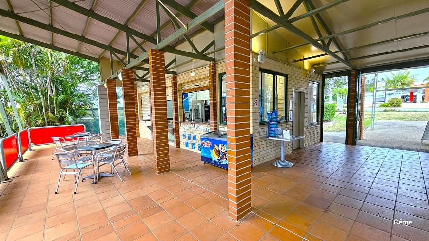 Side angle of the reception and its seating area in front. Separating the reception from the pool are low red barricades with grey steel frames. On the cream brick wall below the reception countertop is ‘GO SWIM’ and several blue graphics. The building has light brown brick foundations, narrow green metal roof beams, and corrugated roofing. The reception area has cream roll-down shutters with light brown accents.