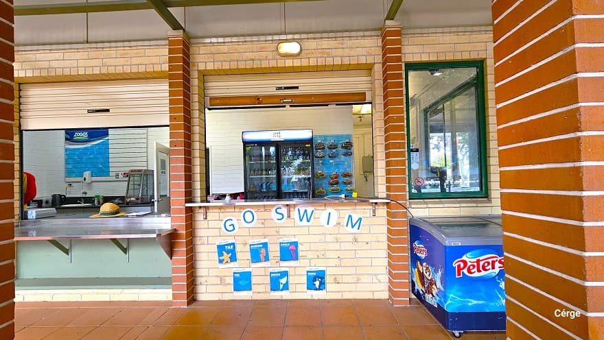 Front of the reception and kiosk at Murrumba Downs Swimming Pool. It has a rectangular glass window, a stainless steel countertop, and cream-colored roll-down shutters with light brown accents for the metal handle for easy access to the shutter. There is a light brown hat on top of the countertop. On the cream brick wall below the reception countertop is ‘GO SWIM’ and several blue graphics. On the right is a blue fridge with ice cream graphic design.