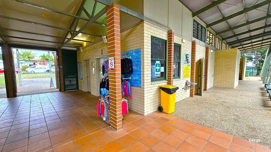 Found opposite the reception at Murrumba Downs Swimming Pool are two white doors accessible along the corridor right after passing the green steel panel Louvre doors. Next to the second white door is the kid's swimwear display. There is a small white ‘No Smoking’ sign on the light brown brick foundation. In front of the glass window with a green cross is a yellow garbage bin with wheels. Open windows near the ceiling provide ventilation for the male and female change rooms.