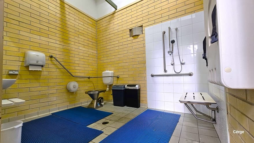 Inside the accessible bathroom at Murrumba Downs Swimming Pool. Near the door is a white adult change table with blue straps. Next to it is the shower section which is highlighted by the white tile wall that contains the shower head, grab bars, and a wall-mounted white shower seat. On the grey tile floor are blue waterproof mats. At the back is the toilet, two black garbage bins, and a grab bar. On the left wall is a white tissue dispenser, a wash basin, and a white pail.
