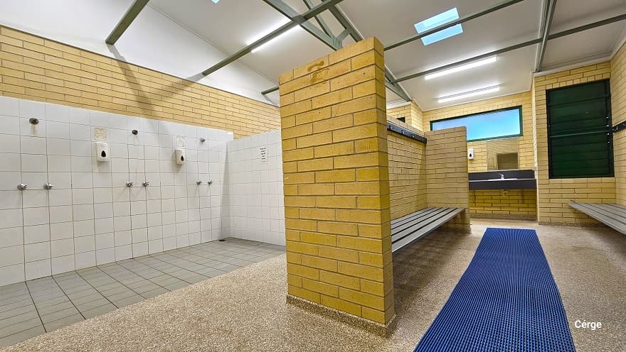 Shower section at Murrumba Downs Swimming Pool has a white tile wall and grey tile floor. A light brown brick wall separates the shower from the waiting area and has wall wall-mounted grey bench and a granite tile floor with a long blue waterproof floor mat at the center. There is a dark wash area towards the exit with a wash basin and a liquid hand soap dispenser. The shower room has a white ceiling with narrow green metal roof beams.