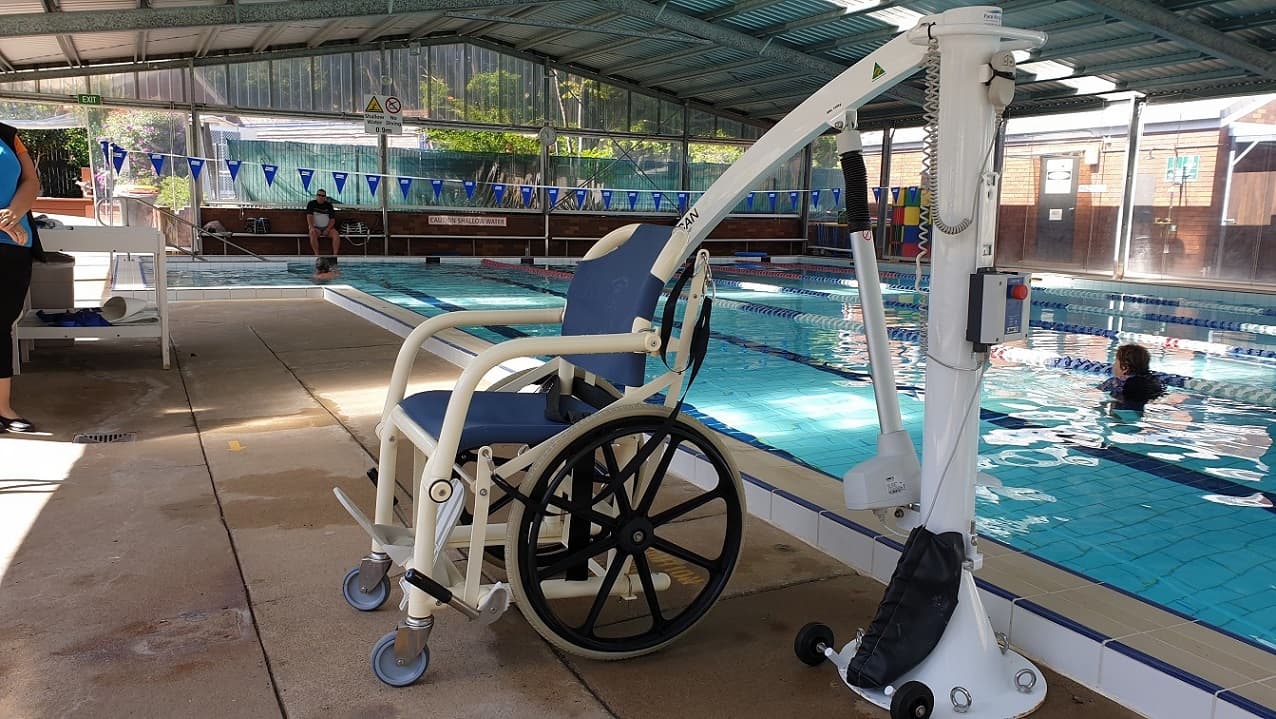 Entry to the 25m Indoor pool includes a pool hoist. A white framed aquatic chair sits attached to a pool hoist to showcase inclusion for mobility disability.