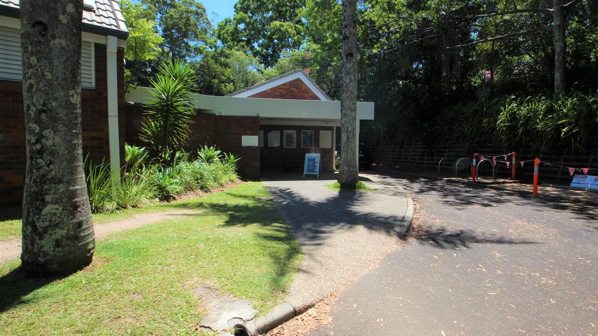 Parking in the accessible car spaces directly in front of the Nambour Aquatic Centre shows a step free entrance toward the low set brown brick building. The venue is set amongst dense foliage creating significand shading.