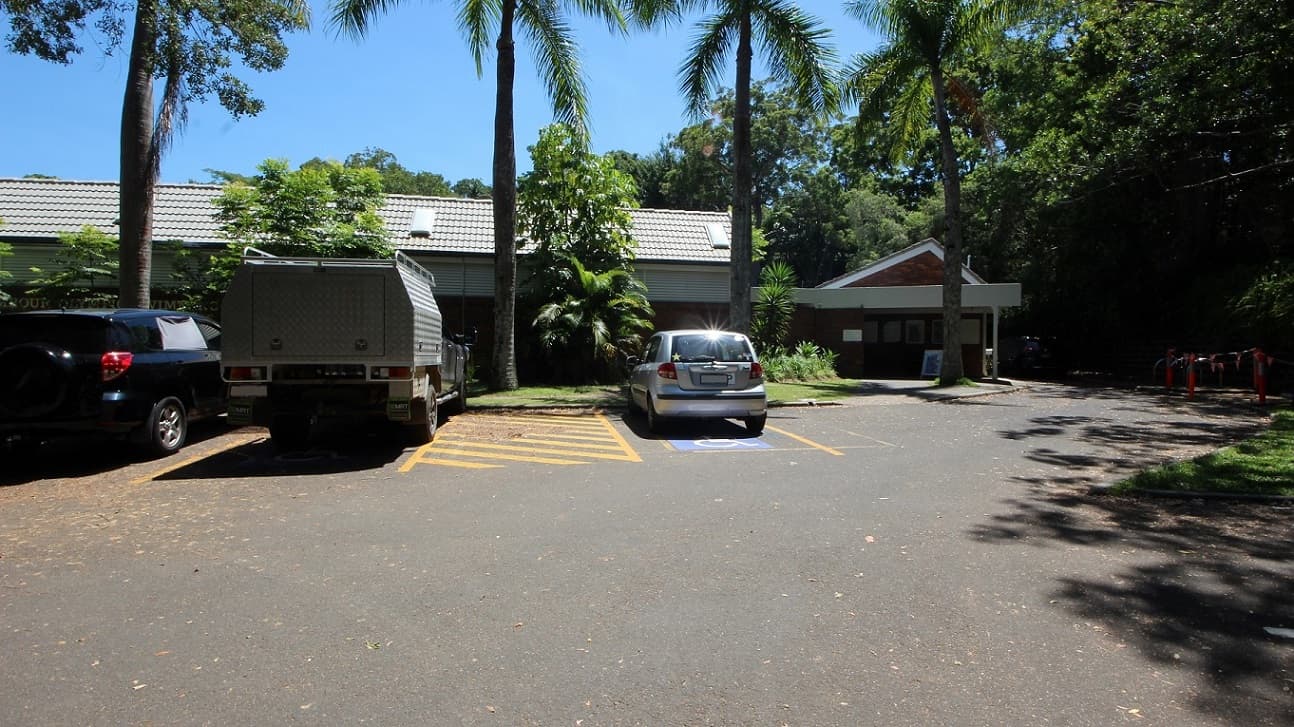 Parking in the accessible car spaces directly in front of the Nambour Aquatic Centre shows a step free entrance toward the low set brown brick building. The venue is set amongst dense foliage creating significand shading.
