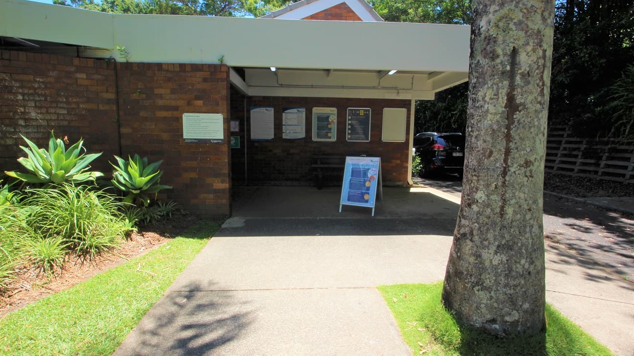 The entrance to the Nambour Aquatic Centre is a step free entrance toward the low set brown brick building. The venue is set amongst dense foliage creating significant shading.