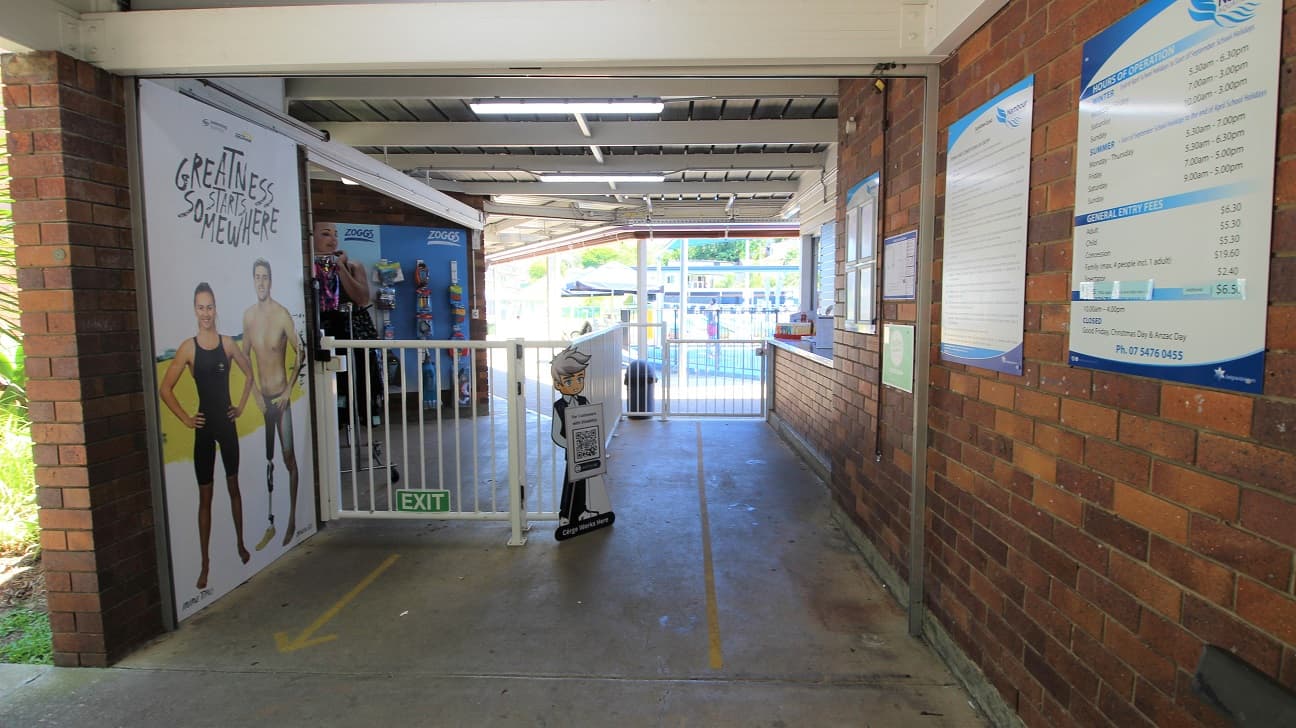 Entrance to the Nambour Aquatic Centre is a passageway design attaching to the reception counter. Entry is via the right and exit via the left. The concrete path and brown brick walls are combined with a white gate to manage the flow of customers.