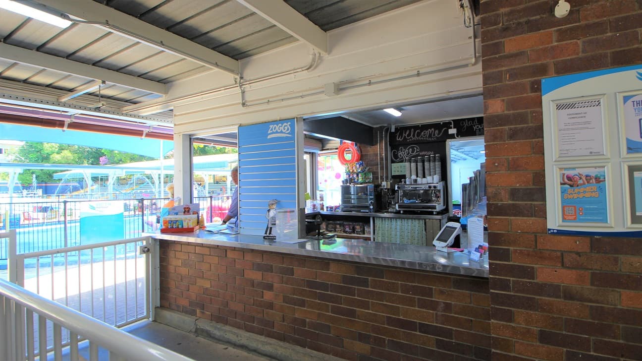 Entrance to the Nambour Aquatic Centre is a passageway design attaching to the reception counter. Entry is via the right and exit via the left. The concrete path and brown brick walls are combined with a white gate to manage the flow of customers.