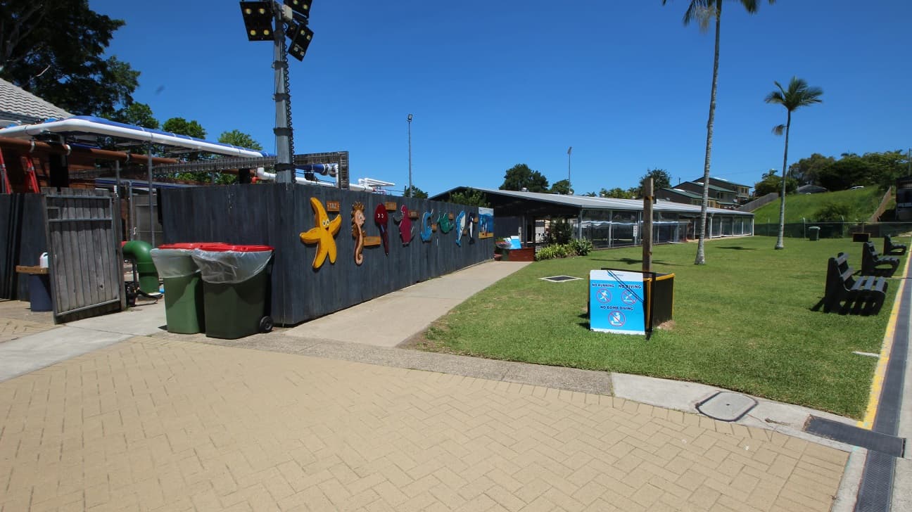 Access to the 25m Indoor Pool is down a slight step free ramp from the main 50m outdoor pool. The concrete path is set next to a 2m dark blue fence on one side and a wide open grassed area to the right.
