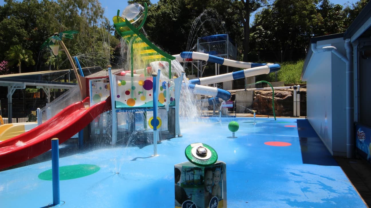 The Nambour Aquatic Centre Splash Park. The aqua blue flooring is contrasted with the red slides and yellow and green equipment. A lot of water is splashing around the equipment. A standard pool fence bounds the splash park from the remainder of the Nambour Aquatic Centre.