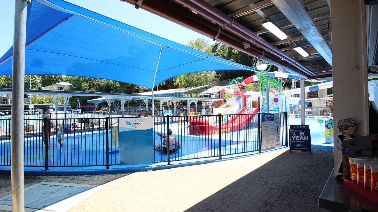 The Nambour Aquatic Centre Splash Park. The aqua blue flooring is contrasted with the red slides and yellow and green equipment. A lot of water is splashing around the equipment.