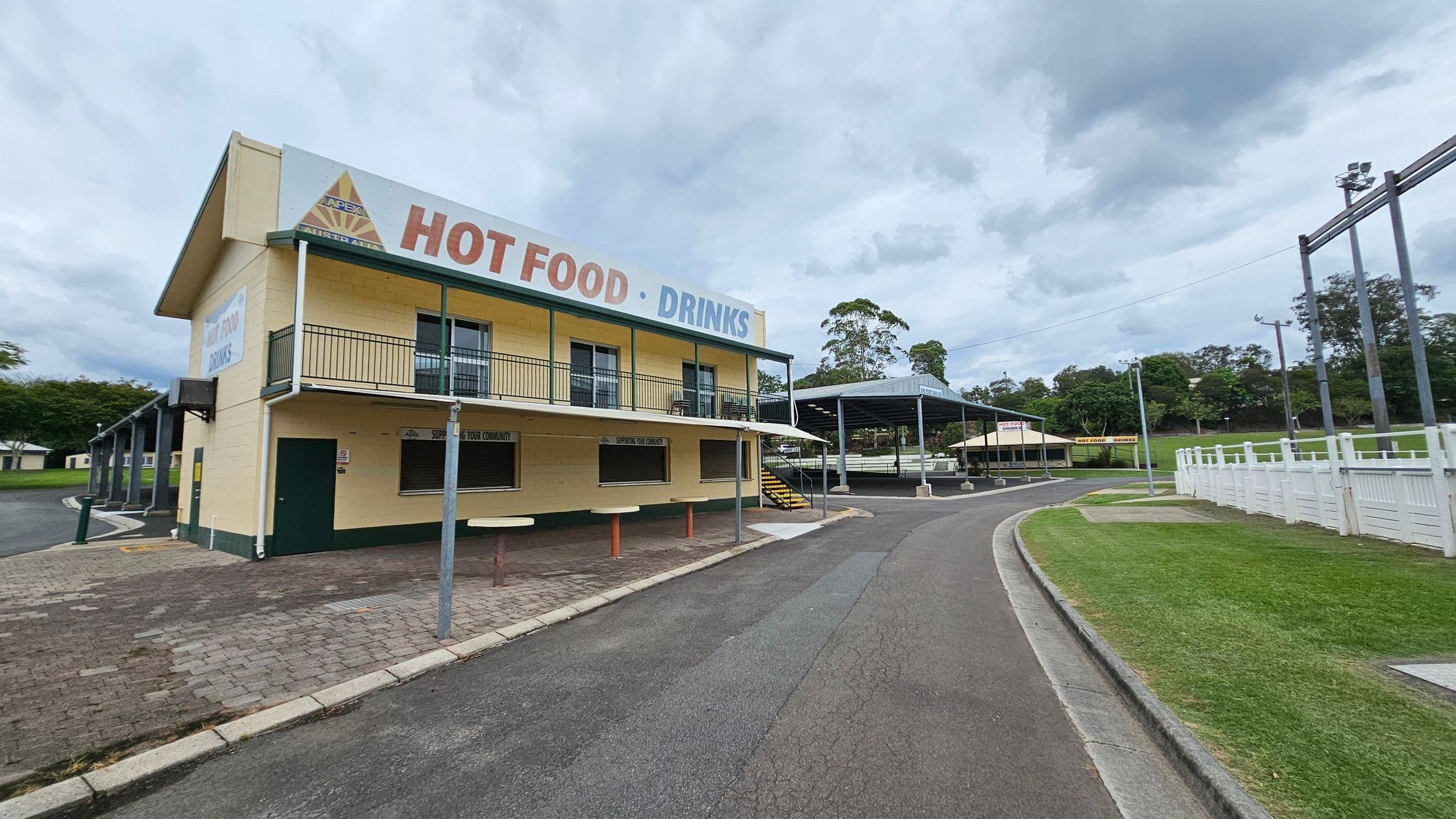 A two-storey yellow building with green trim on the left, featuring a sign that reads "HOT FOOD · DRINKS". It has a balcony and a service window on the ground floor. A paved path leads to a covered seating area with a metal roof in the centre. On the right, there's a curved road bordered by a white fence and a grassy area. Trees and additional structures are visible in the background.