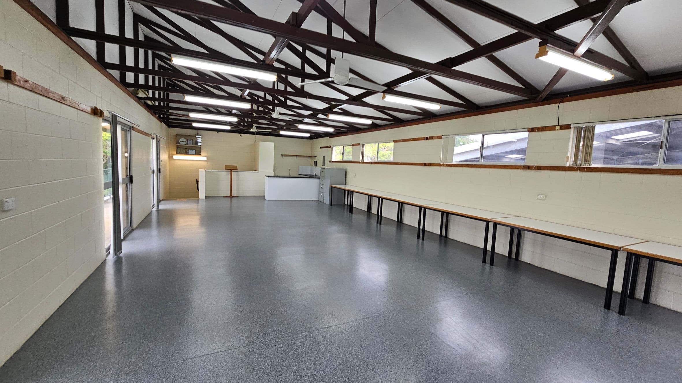 A spacious room with a grey speckled floor and a high ceiling featuring exposed beams. On the left, there are large glass doors. The right side has a long row of tables against the wall, beneath several windows. At the far end, there is a small counter area with a few cabinets. Fluorescent lights are mounted on the ceiling, providing bright illumination.