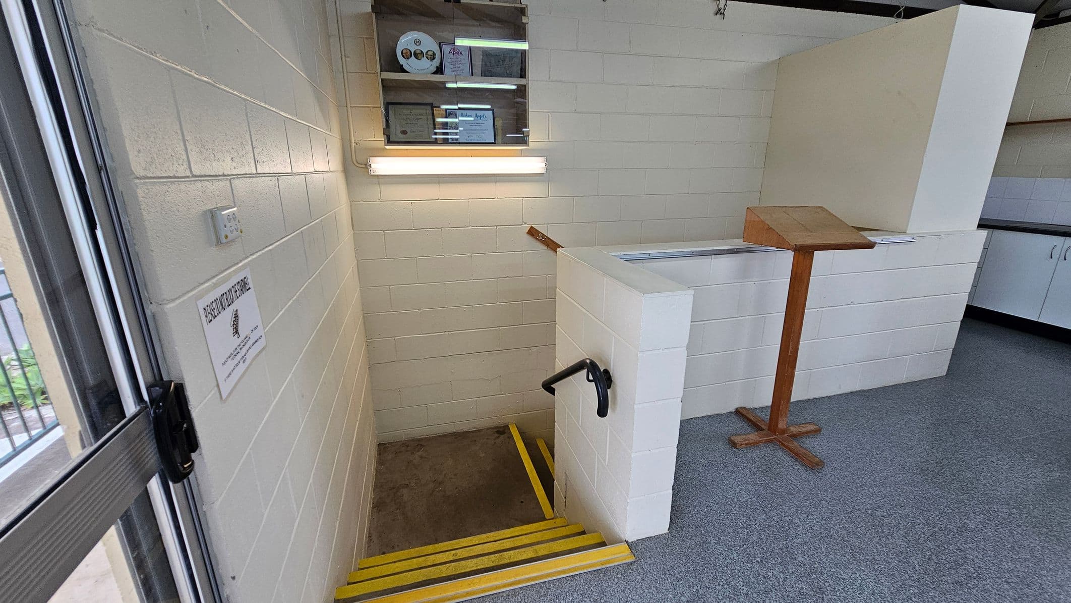 A small stairwell with yellow-edged steps descends into a lower level. On the left, a white brick wall features a sign and a light switch. Above the stairs, a wall-mounted display case holds certificates and a circular item. On the right, a wooden lectern stands on a grey carpeted floor. The ceiling is not visible.