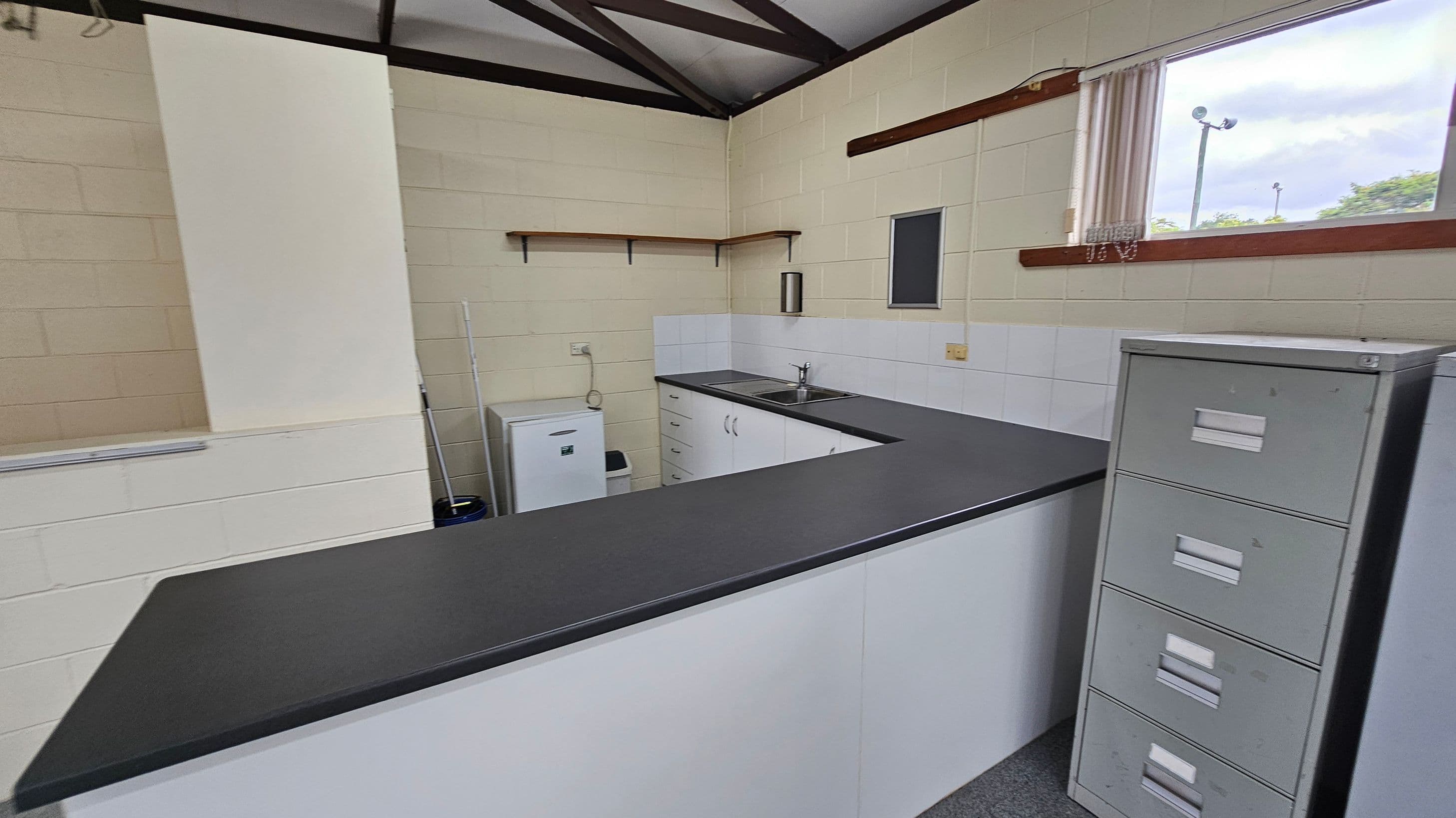 A small kitchen area with a U-shaped black countertop. On the left, there's a white mini fridge and a mop. The back wall has a sink with a double basin, white cabinets, and a small shelf above. On the right, a window with vertical blinds lets in natural light. A grey filing cabinet stands next to the counter. The walls are light green with white tiles around the sink area. The ceiling has exposed beams.