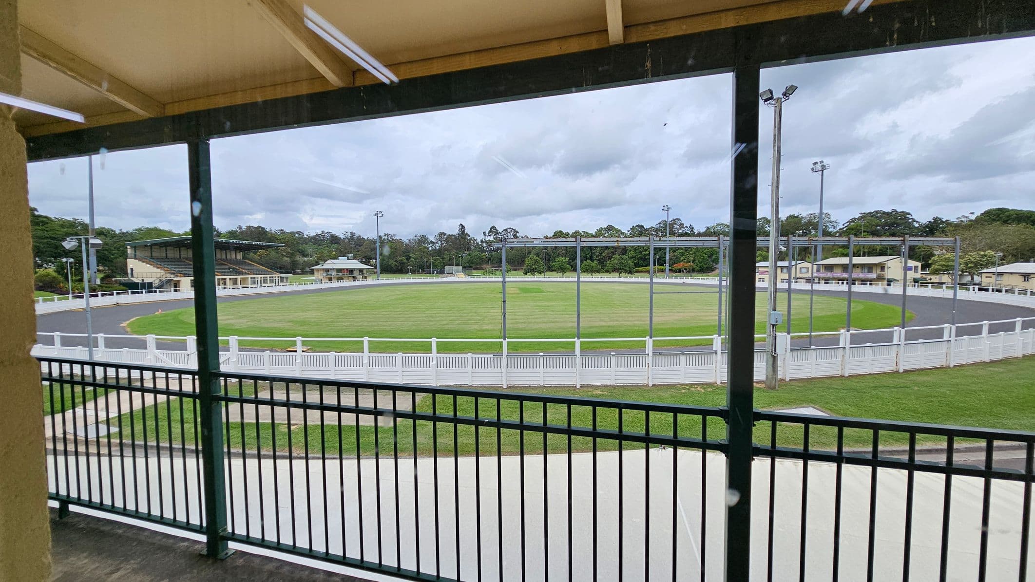 A view from a covered balcony overlooking a circular sports field. The foreground features a black metal railing. Beyond the railing, there's a grassy field encircled by a paved track and a white fence. In the background, there are several buildings, including a grandstand on the left and smaller structures on the right. Tall floodlights are positioned around the field, and trees are visible in the distance. The ceiling of the balcony is wooden with visible beams.