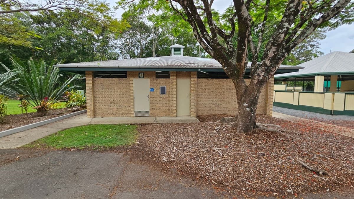 A single-storey brick building with two closed doors in the centre and right. On the left, a concrete path curves alongside a garden with green foliage and small shrubs. A large tree with an uneven bark trunk is in the foreground to the right, surrounded by woodchip ground cover. In the background, another structure with cream walls and a green roof is partially visible. The floor consists of concrete, tarmac, and soil. The ceiling of the visible building is metal roofing.