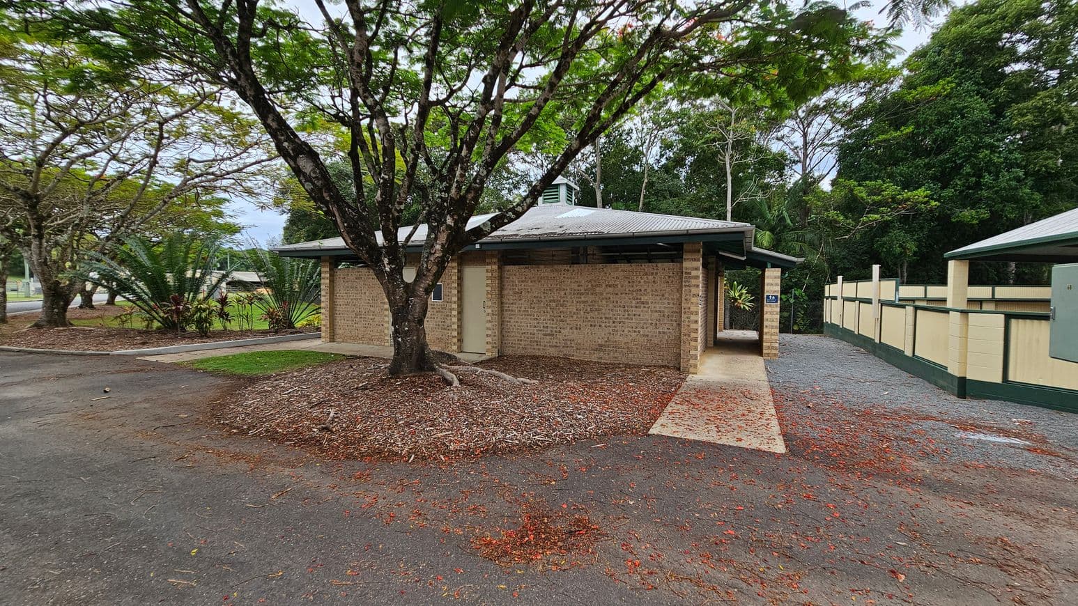 A brick building stands in the centre, with a metal roof and an open passage on the right. On the left, a large tree with a wide, textured trunk is surrounded by woodchip ground cover. On the right, a paved walkway runs alongside a cream and green fence. The ground is a mix of tarmac, concrete, and scattered red leaves. In the background, tall trees and foliage create a dense green backdrop.