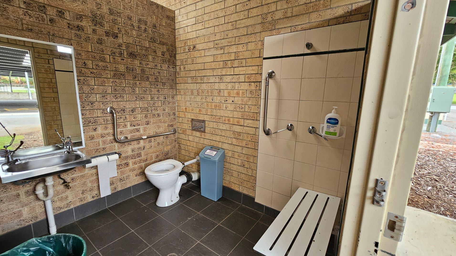 A public restroom with beige brick walls and dark tiled flooring. On the left, a stainless steel sink with a mirror above it and a mounted toilet roll holder beneath. In the middle, a white toilet with a grab bar on the wall behind and a blue sanitary bin to the right. On the right, a tiled wall with attached grab bars, a bottle of soap, and a white slatted bench extending below. The ceiling is cream-coloured with visible piping.