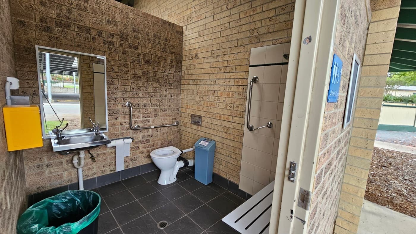 A disabled-access toilet with tan brick walls and dark grey tiled flooring. On the left, a stainless steel sink with a mirror above, a yellow soap dispenser, and a green bin below. In the middle, a white toilet with metal grab rails and a blue sanitary bin beside it. On the right, a tiled shower area with grab bars and a horizontal white bench. The ceiling is light-coloured with exposed piping. The doorway on the right opens to an outdoor area with bark mulch and a green-roofed structure.