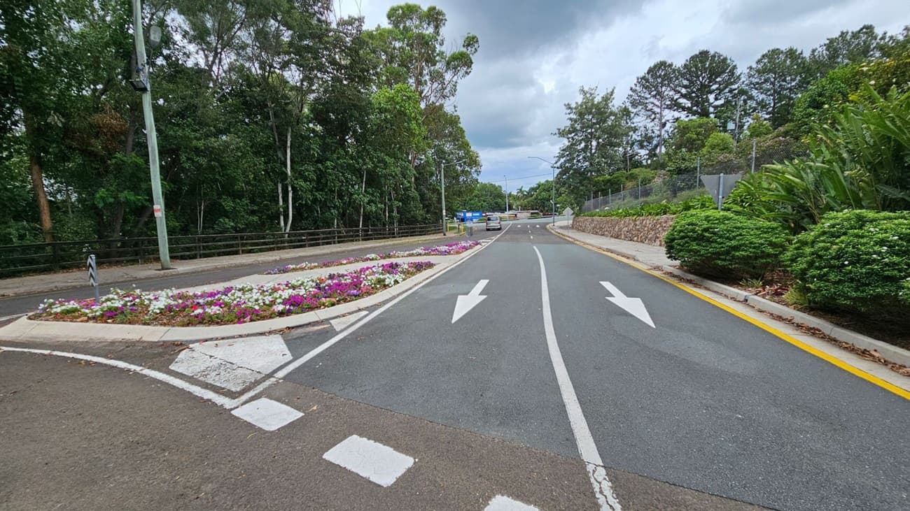 A road curves to the left with white directional arrows painted on it. On the right, a large tree with sprawling roots stands next to a blue sign that reads "Welcome to Nambour Showgrounds." In the background, there is a small building with a light-coloured exterior and a gate. The area is surrounded by lush greenery and trees. The ground is covered with fallen leaves.