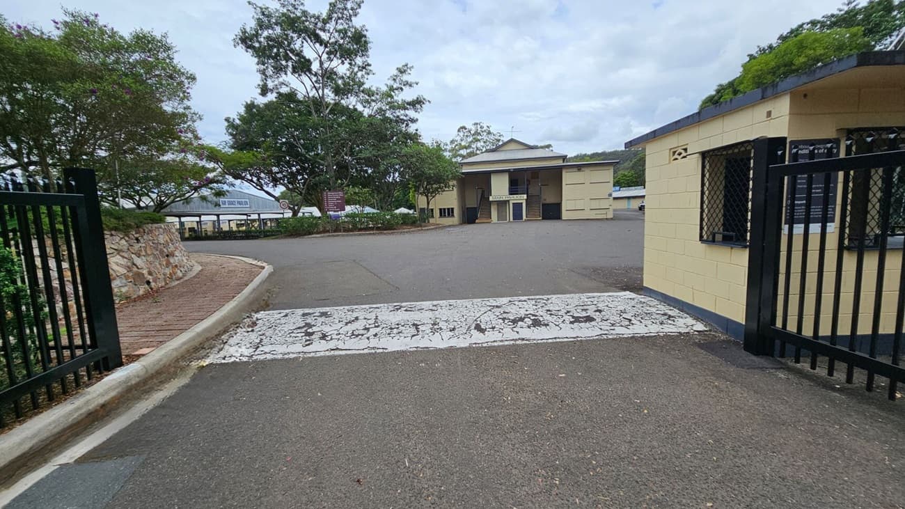A paved road leads to a building with a sign reading "Main Pavilion" in the centre. On the left, there's a stone wall with trees and a path curving alongside it. On the right, a small yellow building with barred windows is adjacent to a black metal gate. The ground is marked with a worn white pedestrian crossing. Trees and additional structures are visible in the background.