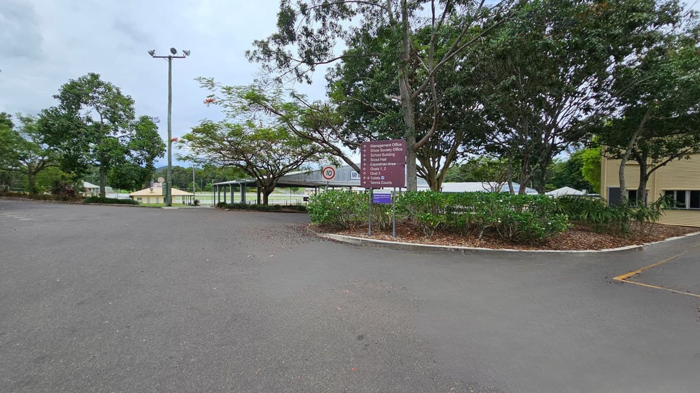 A two-storey building labelled "Main Pavilion" is in the centre, with a staircase leading to the entrance. The building is light-coloured with a gabled roof. On the left, there are trees and shrubs lining a path. On the right, more dense trees and several green bins are visible. The foreground features a large, empty paved area with some scattered leaves and a marked parking space.