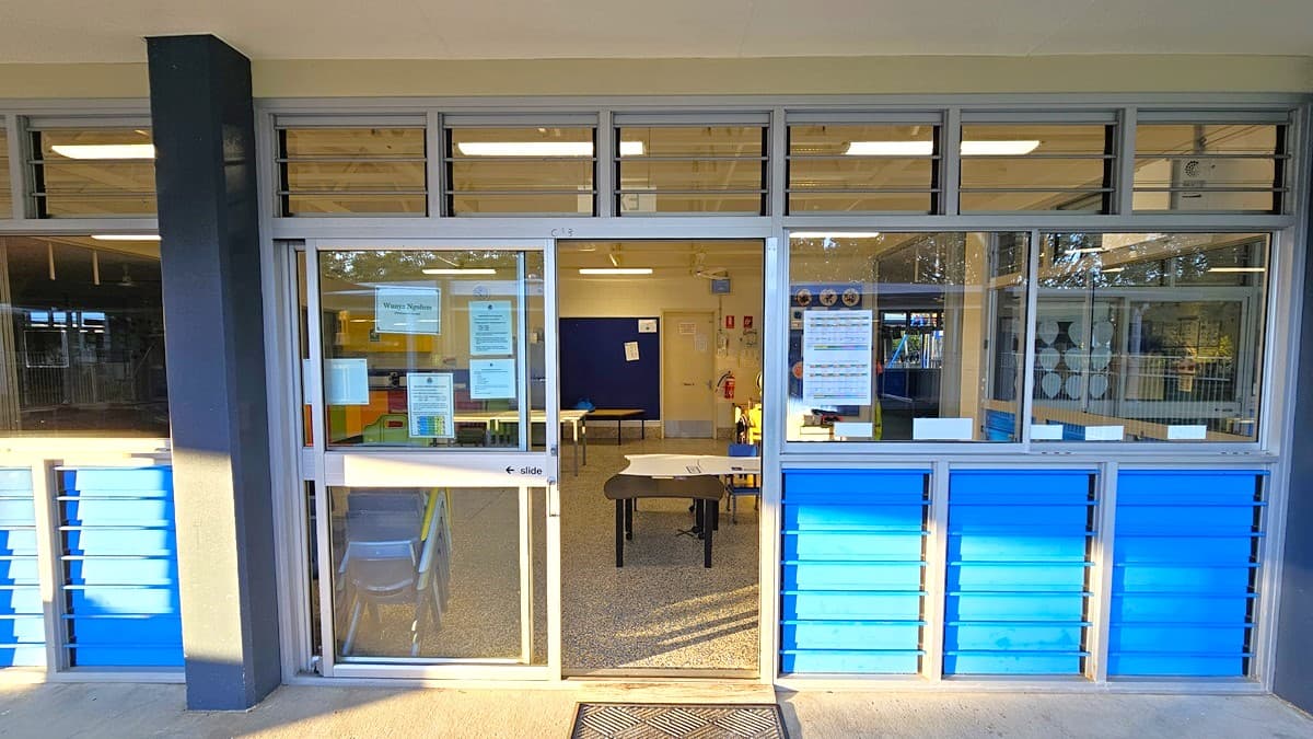 On the left, there is a black column next to a glass door with notices and a sign reading "Office." On the right, there are large glass windows with blue panels below them. In the middle, through the glass door, there is an office with a desk, chairs, and various notices on the walls. The floor inside is covered in grey carpet tiles, and the ceiling is white with exposed beams and fluorescent lighting. The exterior floor is concrete.