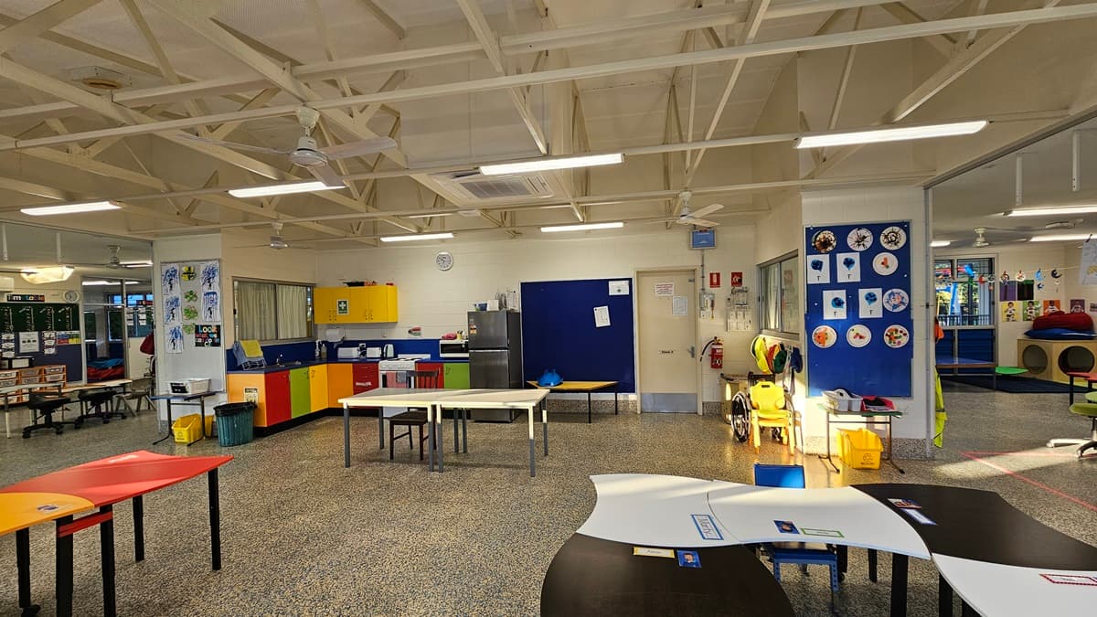 On the left, there are tables with colourful tops, a yellow and red cabinet, and a wall with children's artwork. On the right, there are more tables, a blue wall with circular art pieces, and a yellow walker. In the middle, there is a door with a clock above it, leading to a room with a refrigerator and storage cabinets. The floor is speckled grey, and the ceiling is white with exposed beams, fluorescent lights, and ceiling fans.