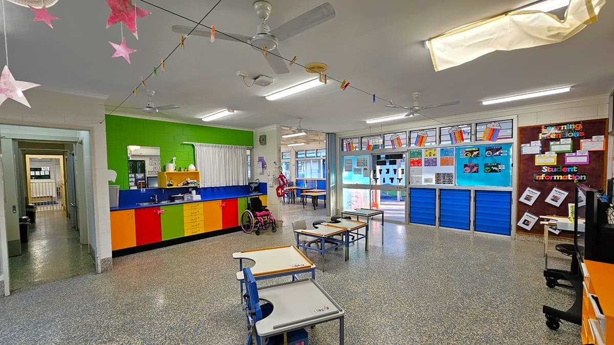 On the left, there is a green wall with a window and a counter below it, featuring colourful cabinets and a pink wheelchair. On the right, there is a blue wall with bulletin boards displaying various papers and a "Student Information" sign. In the middle, there are tables and chairs, with a doorway leading to another room. The floor is speckled grey, and the ceiling is white with fluorescent lights, ceiling fans, and hanging decorations.