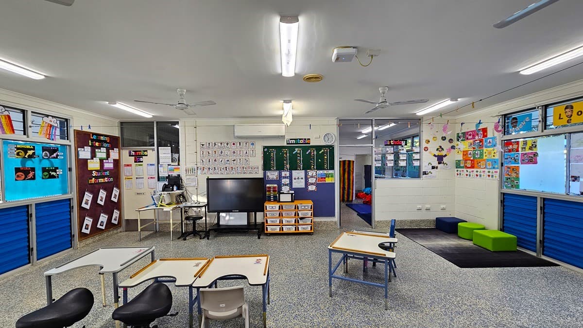 On the left, there are blue storage units below windows, a bulletin board with papers, and a small desk with a computer and a large screen. On the right, there are more blue storage units below windows, colourful artwork on the wall, and green and blue cushioned seats. In the middle, there are tables and chairs, a green chalkboard with educational posters, and a doorway leading to another room. The floor is speckled grey, and the ceiling is white with fluorescent lights and ceiling fans.
