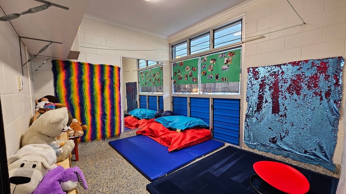On the left, there are shelves filled with stuffed animals and toys, next to a wall covered with a rainbow-striped fabric. On the right, there is a wall with a blue and silver sequined fabric. In the middle, there are blue mats on the floor with red and blue cushions, and windows above with green boards displaying photos. The floor is speckled grey carpet, and the ceiling is white with recessed lighting.