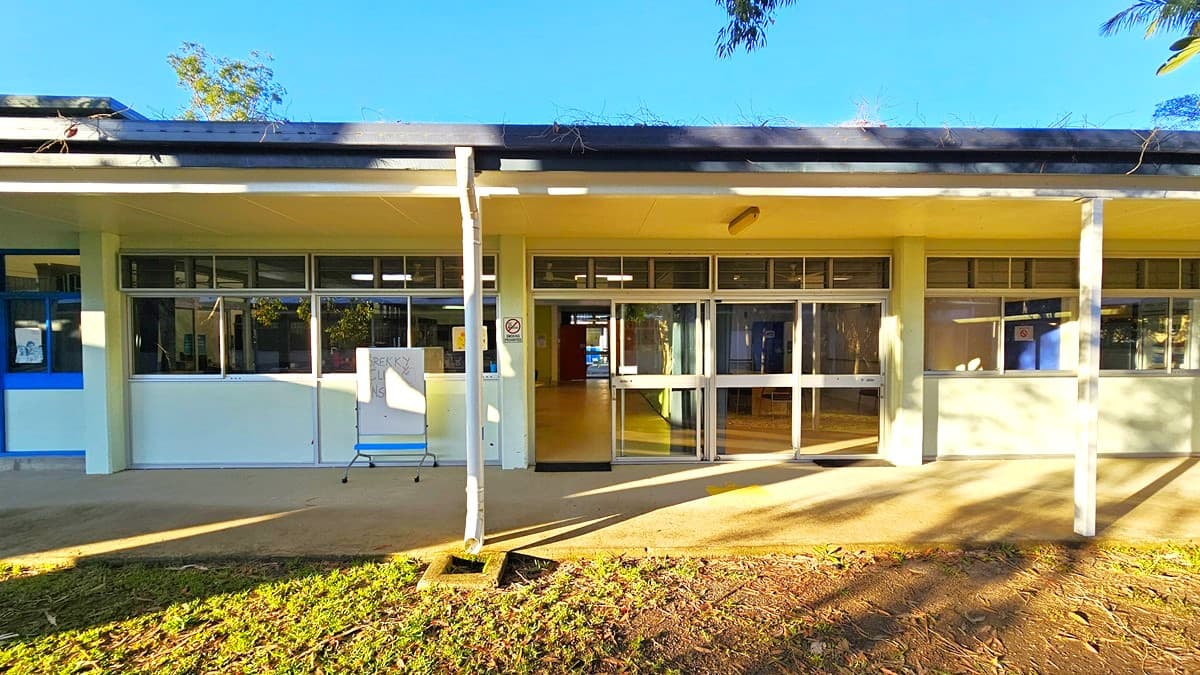 On the left, there is a corridor with windows and blue pillars, leading to a grassy area. On the right, there is a long walkway with a white wall and doors, extending towards another building in the distance. In the middle, there is a corner with a drinking fountain and signs reading "Multi-Purpose Block G" and "D Block." The floor is concrete, and the ceiling is white with fluorescent lights.