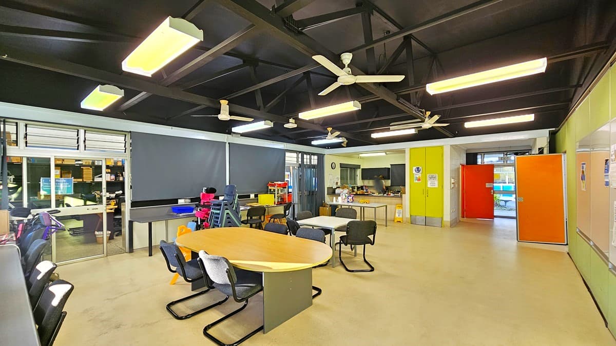 On the left, there are windows with blinds, a black desk with a computer, and a stack of colourful chairs. On the right, there is a green wall with a bulletin board and an orange door. In the middle, there is a round wooden table with black chairs around it. The background features a kitchen area with a sink, cabinets, and a doorway. The floor is light-coloured concrete, and the ceiling is black with exposed beams, fluorescent lights, and ceiling fans.