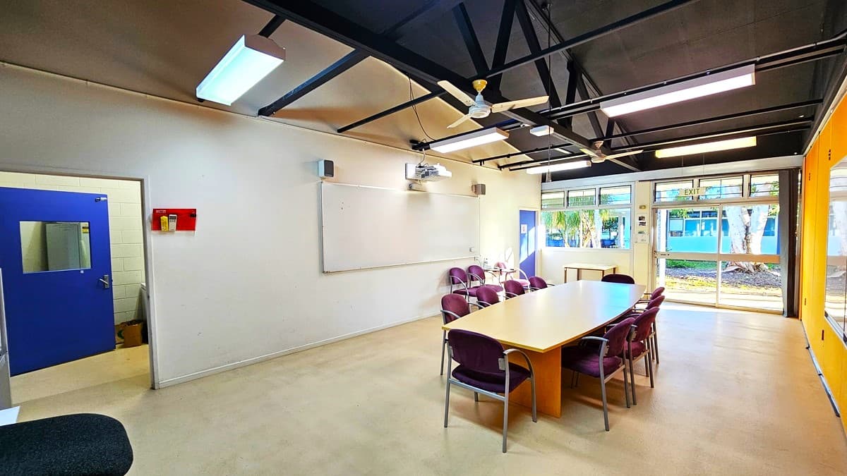 On the left, there is a white wall with a blue door featuring a small window, a red fire alarm box, and a whiteboard with speakers above it. On the right, there are large windows with a view of trees and a building outside, and an orange wall with a blue door. In the middle, there is a long wooden table surrounded by purple chairs. The floor is light-coloured concrete, and the ceiling is black with exposed beams, fluorescent lights, and ceiling fans.