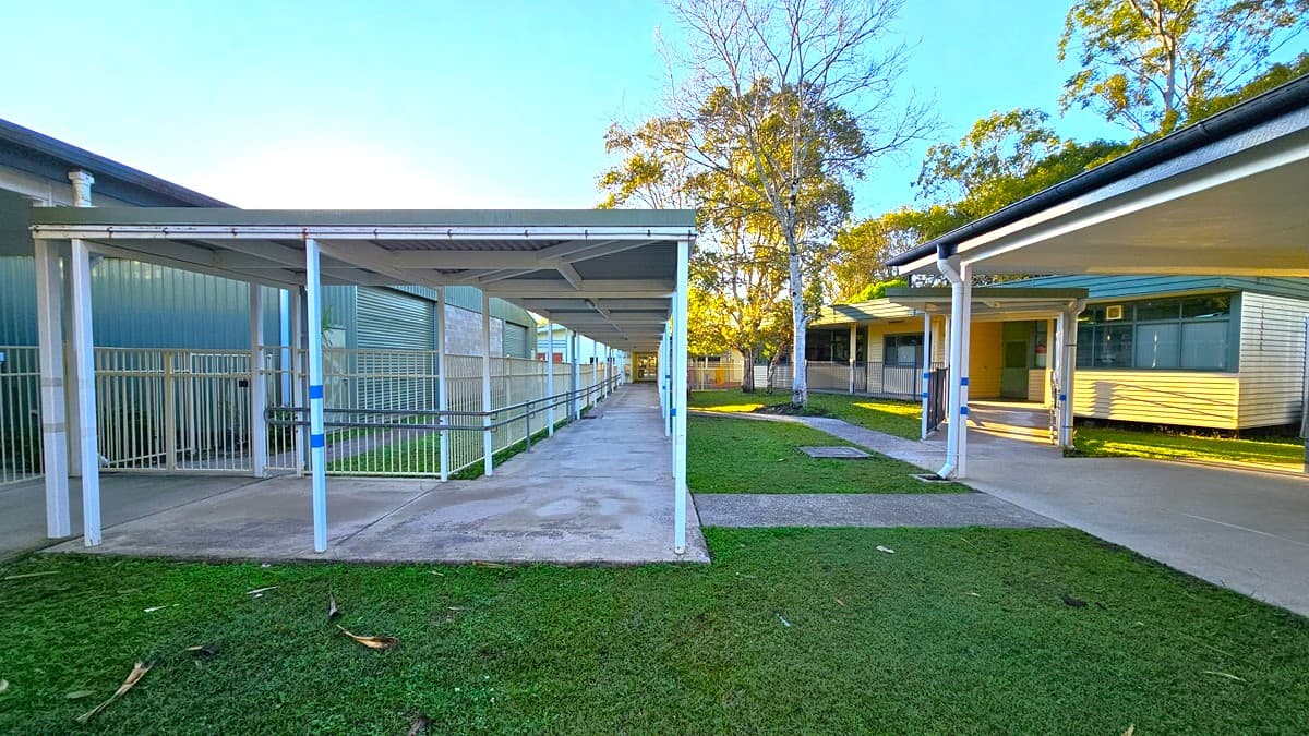On the left, there is a covered walkway with white railings and a green building with vertical panels. On the right, there is another covered walkway leading to a building with large windows and a yellow wall. In the middle, there is a concrete path flanked by grass, leading to a tree and more buildings in the background. The floor is concrete, and the ceiling of the walkways is white with metal supports.