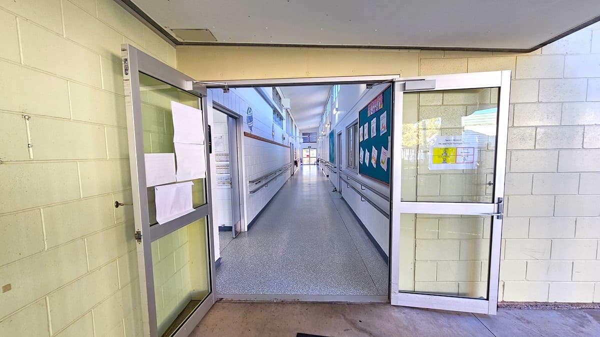 On the left, there is an open glass door with papers taped to it and a beige tiled wall. On the right, there is another open glass door with a green bulletin board displaying various notices and a beige tiled wall. In the middle, there is a long corridor with white walls, handrails, and a speckled grey floor, leading to a distant area. The ceiling is white with fluorescent lights. The floor at the entrance is concrete.