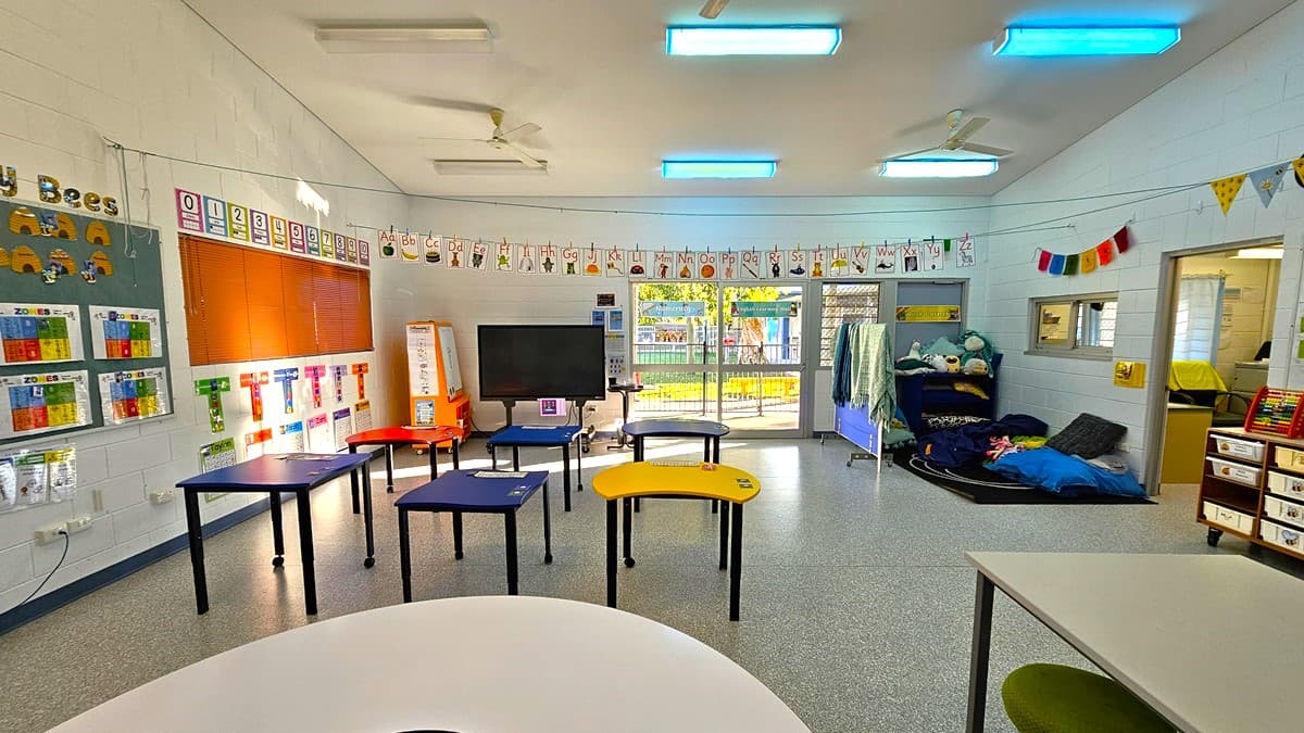 On the left, there is a bulletin board titled "Birthday Bees" with colourful charts and a large orange notice board below it. On the right, there is a small area with a blue mat, cushions, and a shelf with books and toys. In the middle, there are several tables in blue, yellow, and red, arranged in a semi-circle, with a large screen on a stand. The floor is speckled grey, and the ceiling is white with fluorescent lights and ceiling fans.