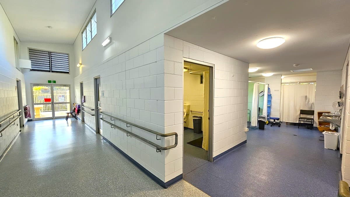 On the left, there is a corridor with white tiled walls, handrails, and windows near the ceiling. At the end of the corridor, there is a glass door leading outside. On the right, there is a room with a blue floor, a white curtain, a sink, and a small table. In the middle, there is an open doorway leading to a room with a toilet. The floor is speckled grey in the corridor and blue in the rooms. The ceiling is white with recessed lighting.