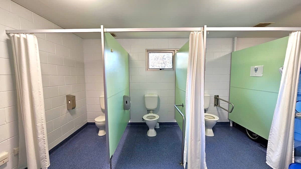On the left, there is a white tiled wall with a toilet cubicle featuring a green partition and a white curtain. In the middle, there is another toilet cubicle with a green partition, a white curtain, and a small window above the toilet. On the right, there is a third toilet cubicle with a green partition, a white curtain, and a grab bar next to the toilet. The floor is blue with a textured surface, and the ceiling is white with recessed lighting and vents.