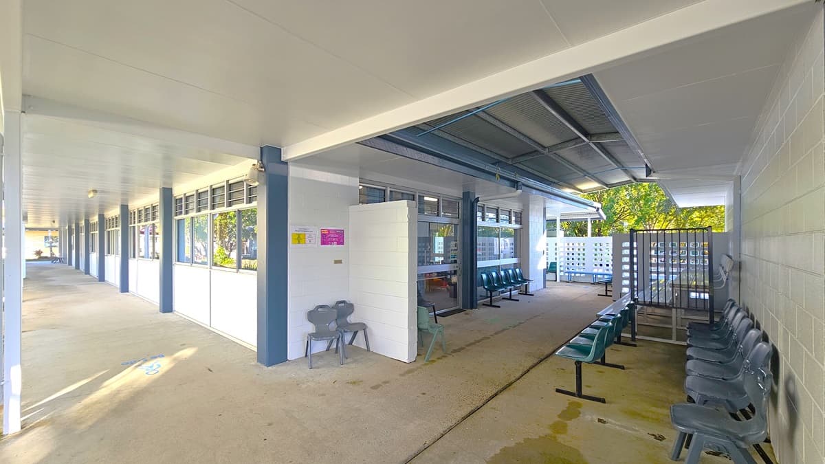 On the left, there is a long covered walkway with white pillars and windows along the wall. In the middle, there is an open area with a white door and a few grey plastic chairs. On the right, there are rows of green plastic chairs facing each other, separated by a narrow walkway. The floor is concrete, and the ceiling is white with recessed lighting. The background includes more windows, a lattice fence, and trees visible through the openings.