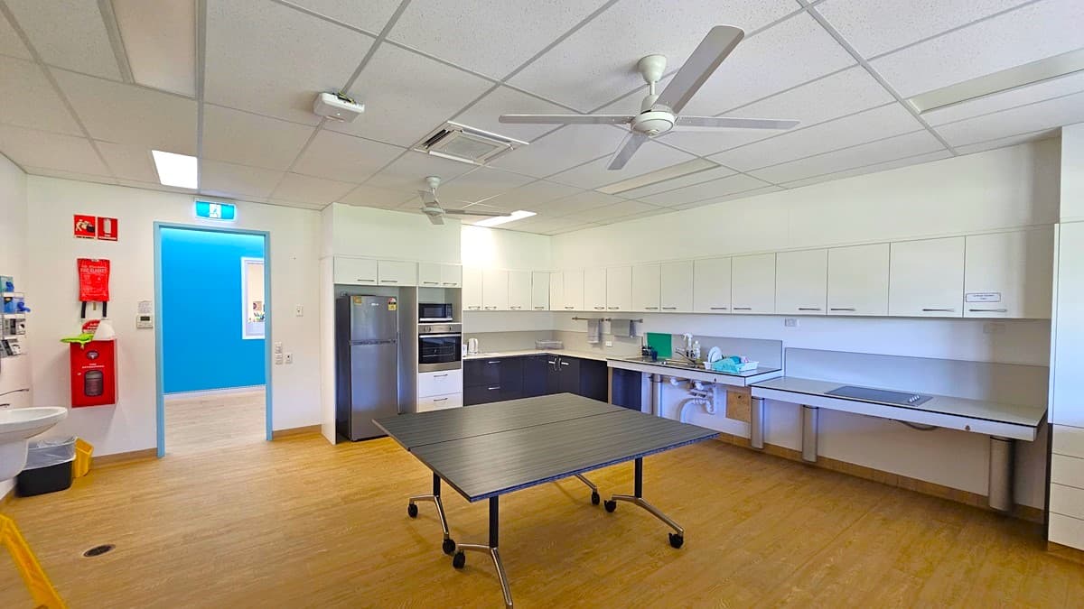 On the left, there is a white door with a blue exit sign above it, a fire extinguisher, and a fire hose reel mounted on the wall. In the middle, there is a black table with wheels on a wooden floor. On the right, there are white cabinets and countertops with a sink, microwave, and various items. The ceiling has white tiles, fluorescent lights, ceiling fans, and an air conditioning vent.