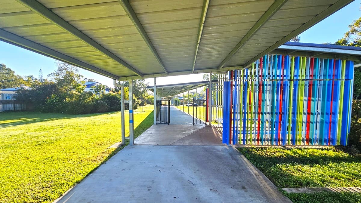 On the left, there is a grassy field with trees and a building in the distance. In the middle, there is a concrete pathway extending straight ahead, covered by a metal roof supported by red and grey poles. On the right, there is another grassy field with a building featuring a covered walkway and windows. The floor is a concrete path, and the ceiling is a corrugated metal roof.