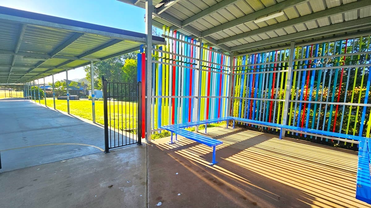 On the left, there is a covered walkway with a metal roof supported by grey poles, leading to a grassy field with trees and buildings in the distance. In the middle, there is a black metal gate and a concrete floor. On the right, there is a seating area with blue benches and colourful vertical stripes on the walls. The floor is concrete, and the ceiling is a corrugated metal roof.