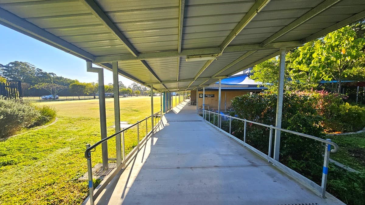 On the left, there is a grassy field with trees and a fence in the distance. In the middle, there is a long concrete walkway with metal railings, covered by a corrugated metal roof supported by grey poles. On the right, there are bushes and a building with a blue roof partially visible. The floor is concrete, and the ceiling is a corrugated metal roof.
