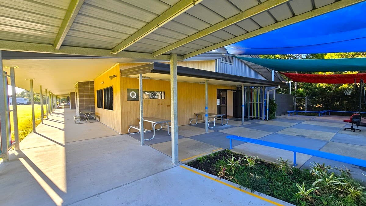 On the left, there is a covered walkway with a metal roof supported by grey poles, leading to a building with beige walls and a sign reading "Q Senior Unit Block 1." In the middle, there are concrete pathways and a few benches. On the right, there is an open area with blue and red shade sails above, a blue bench, and a small garden with plants. The floor is concrete, and the ceiling is a corrugated metal roof.